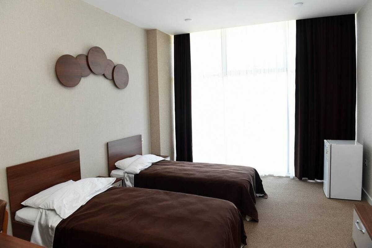 Ett hotellrum med två sängar, ett litet vitt kylskåp och ett fönster med gardiner.