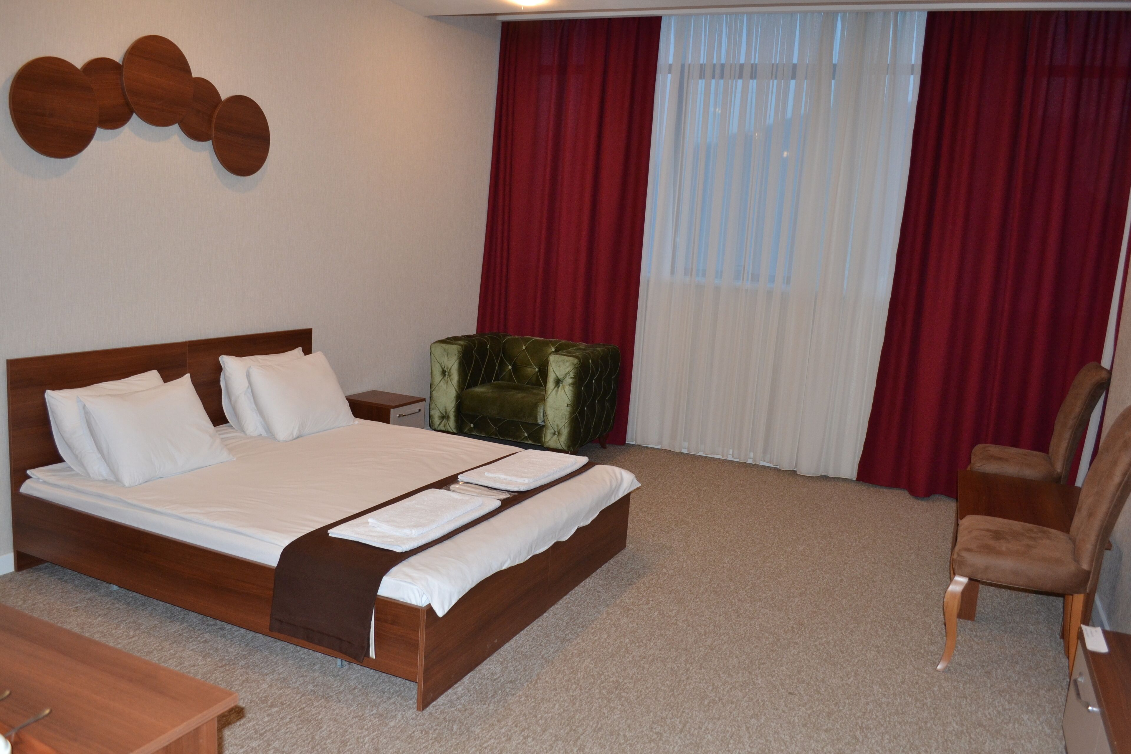 A hotel room with a large bed, a green upholstered chair, a wooden nightstand, and a wooden desk.