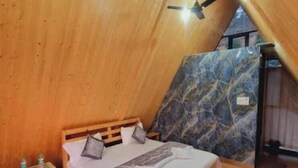 A cozy bedroom with a sloped ceiling, a bed with pillows, a small wooden nightstand, and a purple rug. - Veda Cottage (Barkot)