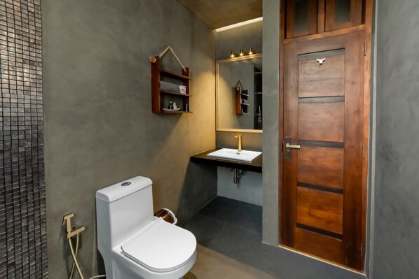 Un baño moderno con inodoro blanco, puerta de madera, espejo y un pequeño estante con artículos de aseo.