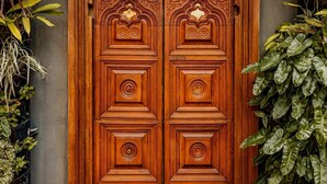 Een houten dubbele deur met ingewikkelde houtsnijwerken, voor een muur met weelderige begroeiing.