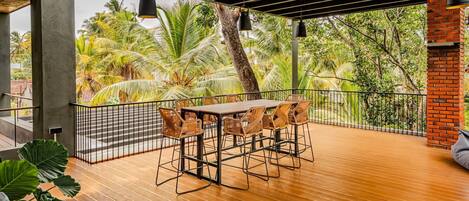 A wooden deck with a table and chairs, surrounded by a metal railing, overlooking a tropical garden with palm trees.