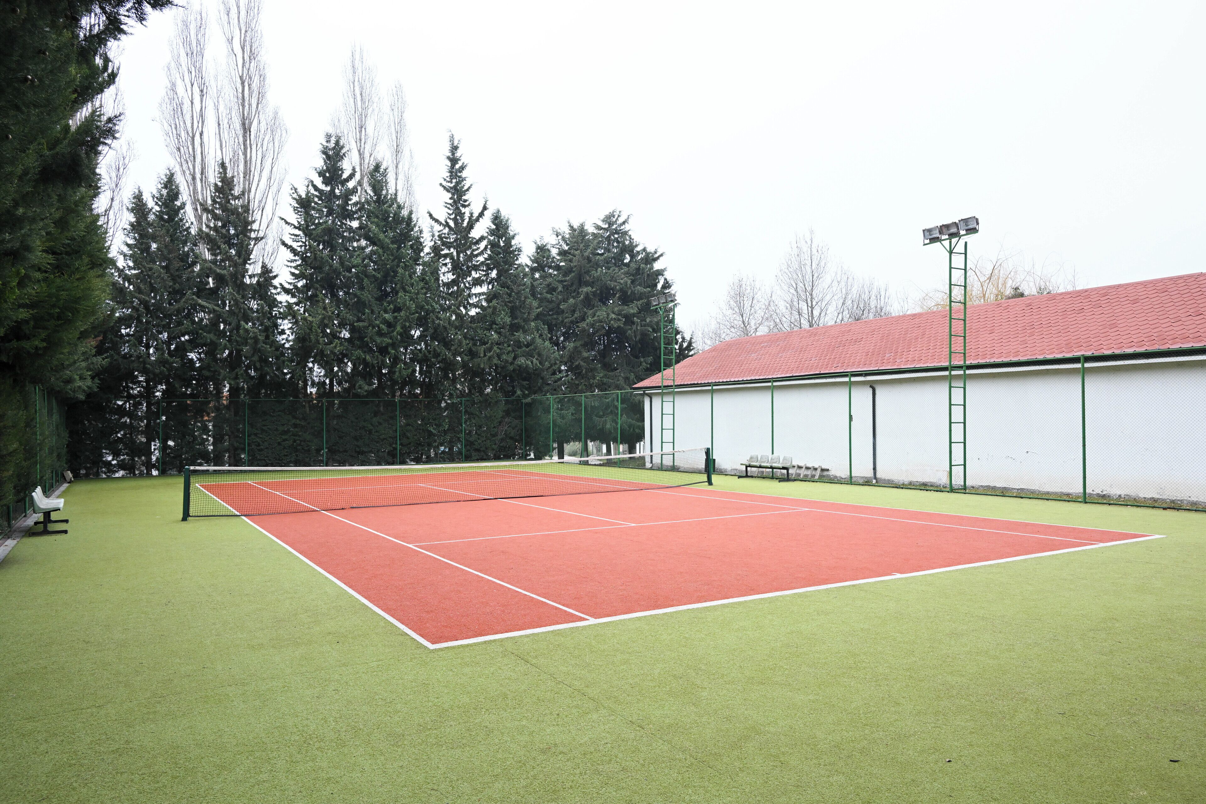 An indoor tennis court with a net, surrounded by trees and a building with a red roof.