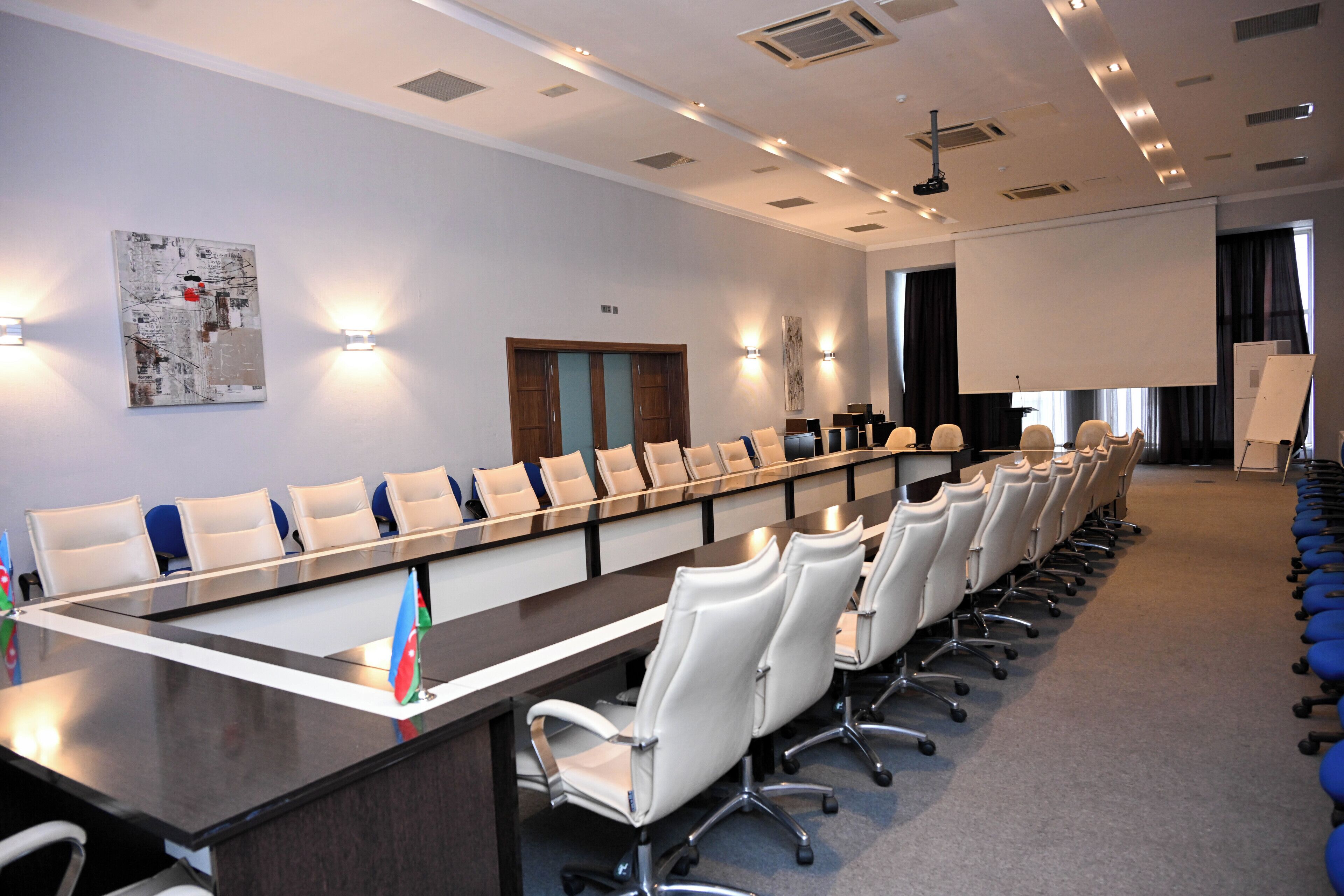 A conference room with a long table, white chairs, and a projector screen.