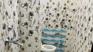 A bathroom with a toilet, a sink, and a blue stool with a bucket. The walls have a floral pattern. - S R HOTEL (Varanasi)
