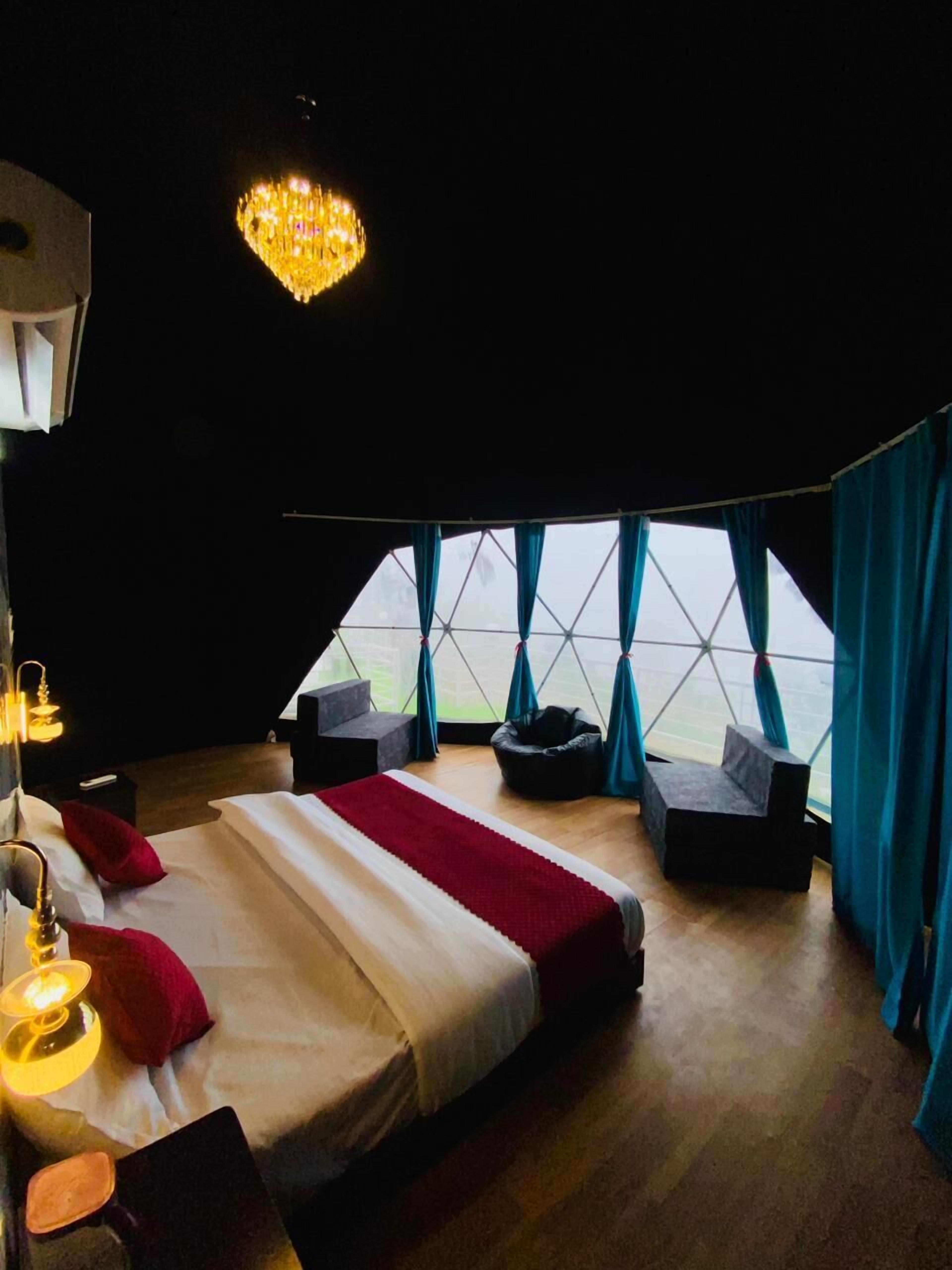 A modern bedroom with a large bed, a chandelier, and a view through a geometric window.