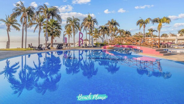 Una piscina con palmeras, sillas de descanso, un puente y un letrero que dice 'Beach Please'.