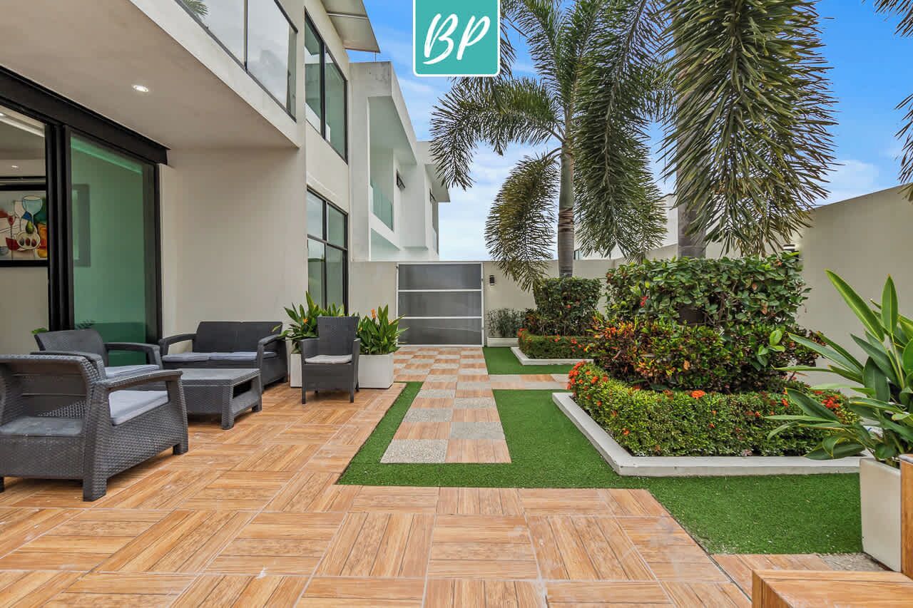 A modern outdoor patio with wicker furniture, a checkered floor, and a landscaped garden with palm trees and shrubs.