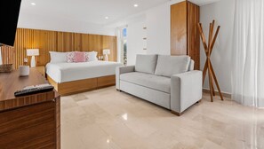 A modern hotel room with a wooden headboard, a white bed, a grey sofa, and a wooden floor. - Beachfront Studio • Rooftop Pool • Ocean Views (Puerto Vallarta)