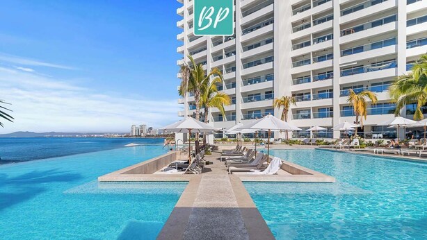 A hotel pool area with lounge chairs and umbrellas, a clear view of the ocean, and a tall building with a BP sign.