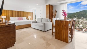 A modern hotel room with a bed, a sofa, a dining table, and a view of a cityscape.