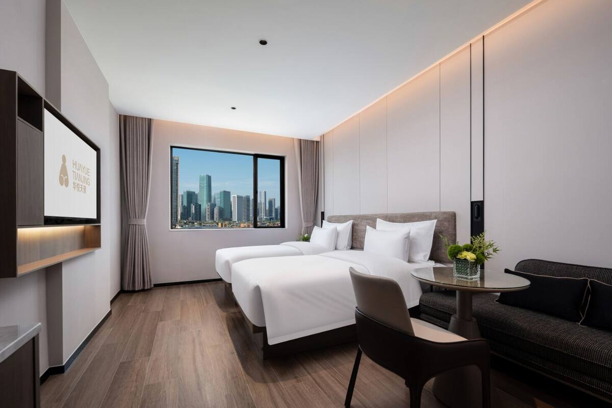 Habitación de hotel moderna con una cama grande, un escritorio y un sofá. Se observa una vista panorámica de la ciudad a través de la ventana.