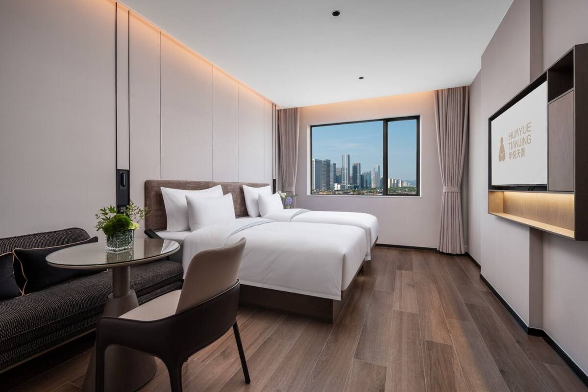 Habitación de hotel moderna con vistas a la ciudad, un sofá, una mesa redonda y una cama con ropa de cama blanca.