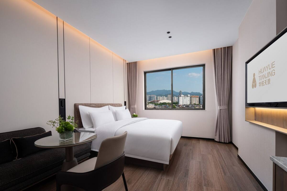 Una habitación de hotel moderna con una cama grande, una zona de estar con mesa y sillas, un amplio ventanal con vista a la ciudad y un televisor montado en la pared.
