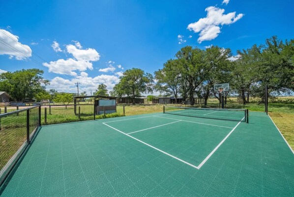 Una cancha de tenis al aire libre con superficie verde, rodeada de árboles y un cielo despejado.