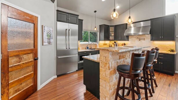 A modern kitchen with dark cabinets, stainless steel appliances, a stone island, and bar stools. - 2 Home Getaway-Pool-Hot Tub-Pickleball Court (Fredericksburg)