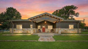 A two-story building with a stone foundation and wooden siding, featuring a front porch with two rocking chairs and a red door. - 2 Home Getaway-Pool-Hot Tub-Pickleball Court (Fredericksburg)