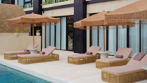 A poolside area with lounge chairs and umbrellas, a clear pool, and a building with large windows and plants on the balconies. - Sunny Samudra  (Pecatu)