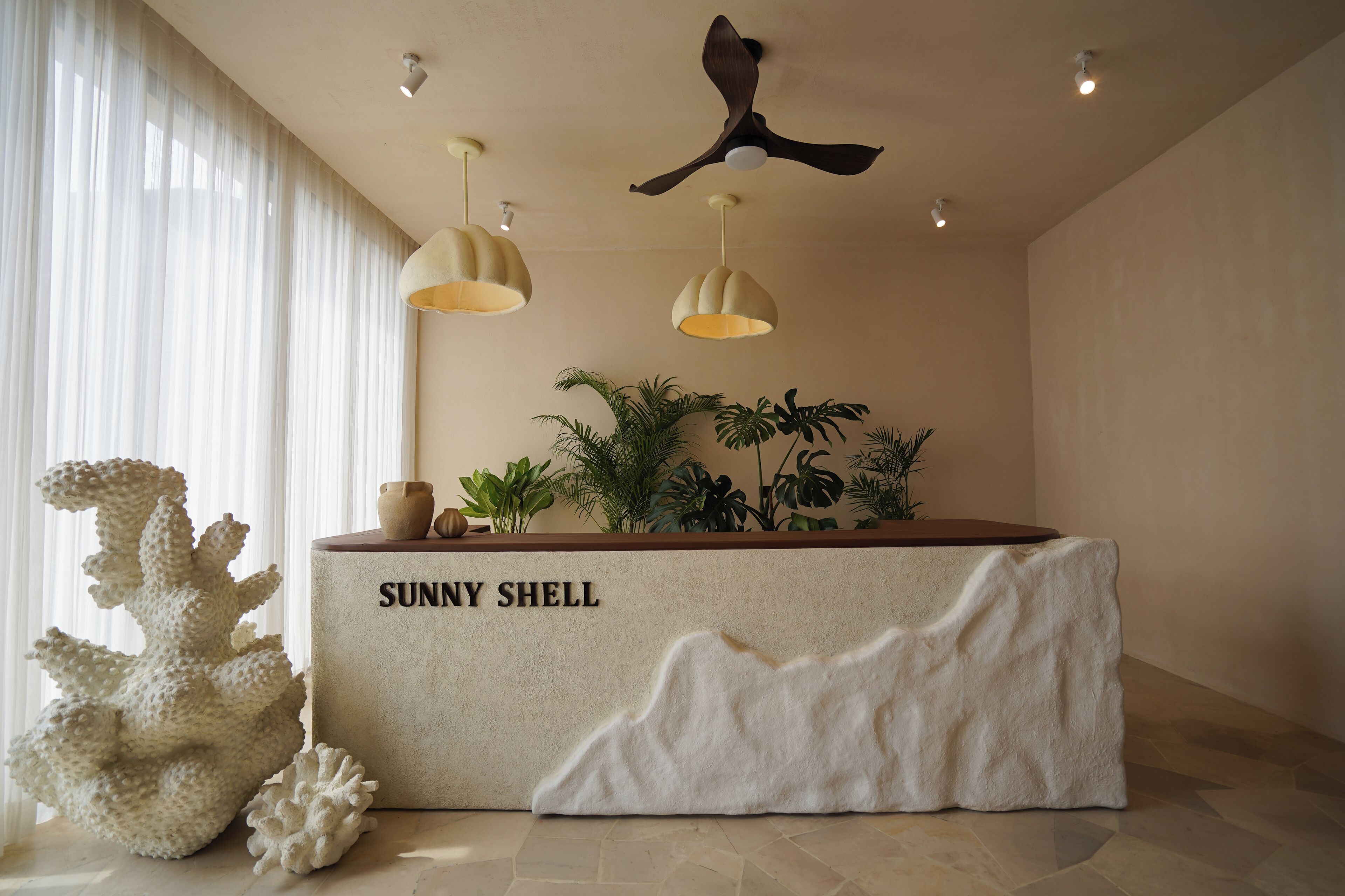 Ein Empfangstresen mit einem Schild 'Sunny Shell', einem Deckenventilator, Pendelleuchten und einer Pflanzengestaltung.