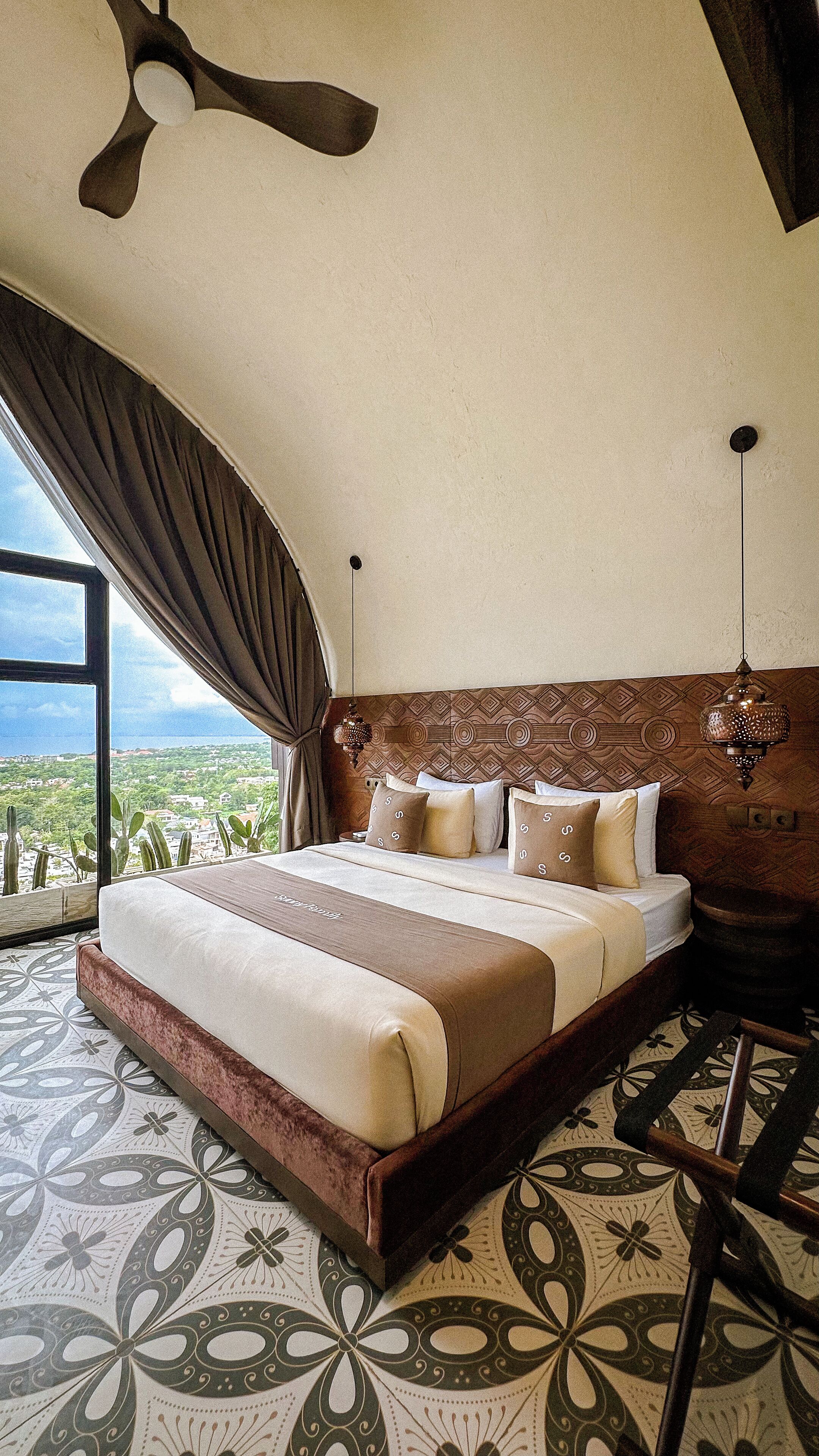 Un'ampia camera da letto con un letto grande, una ventola a soffitto, vista sul paesaggio e un pavimento decorato.