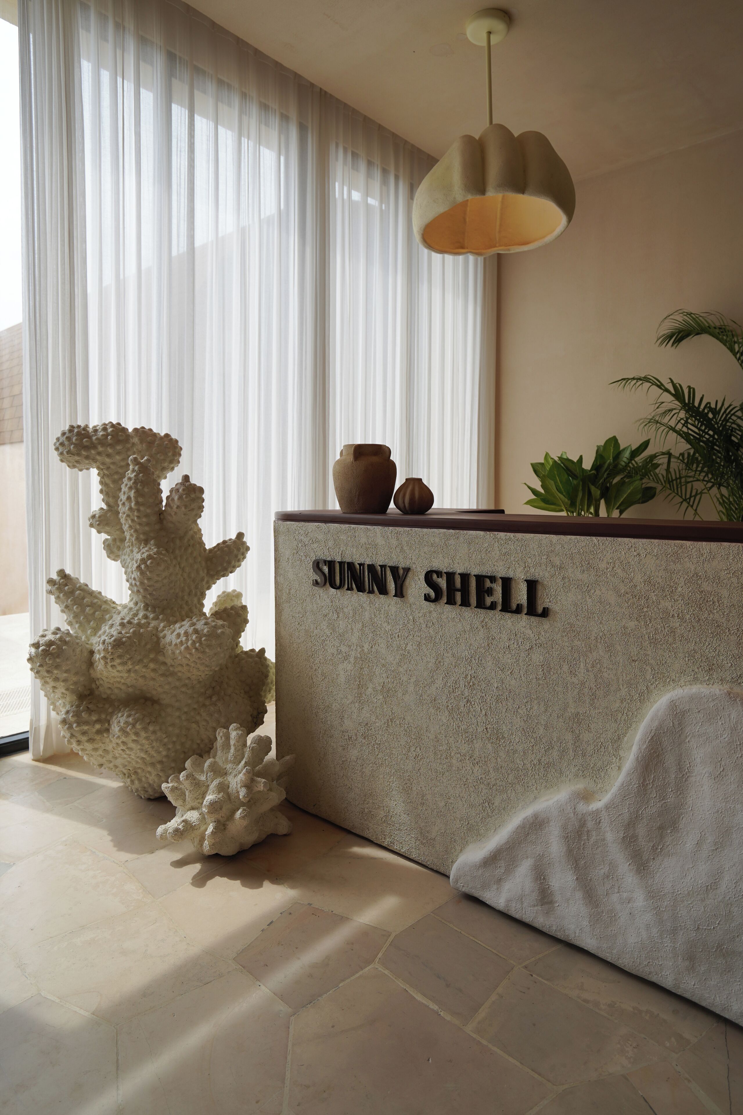 Een receptiebalie met de tekst 'Sunny Shell', een gestructureerde wand, een hangende pendelarmatuur en een grote witte koraal sculptuur.