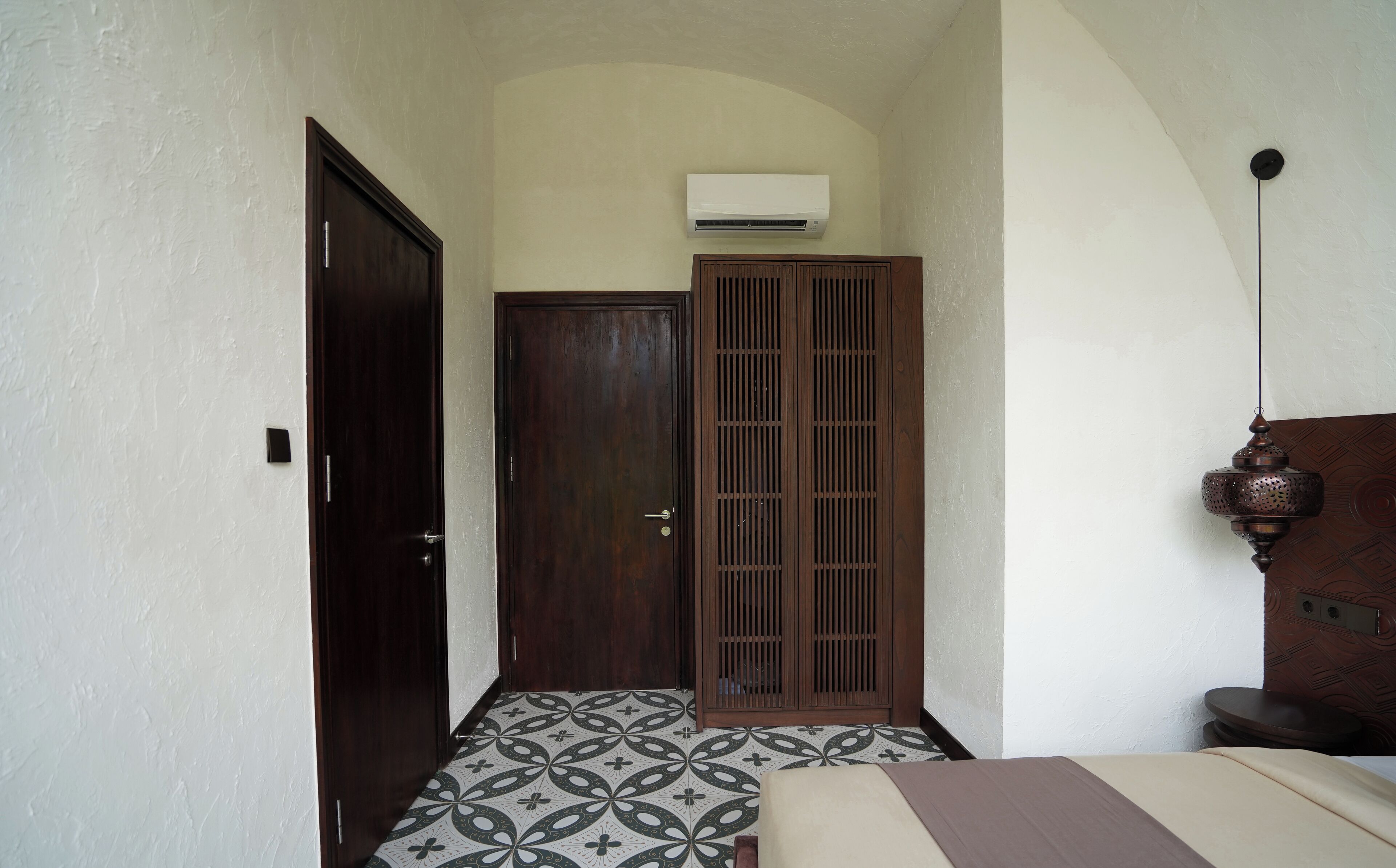 A narrow hallway with a patterned floor, a wooden door, a wardrobe, and an air conditioning unit mounted on the wall.
