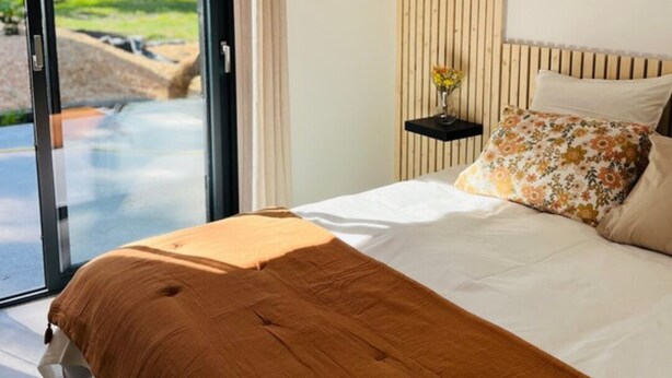 Une chambre dotée d’un grand lit, d’une tête de lit en bois, d’une table de chevet avec un vase, et d’une porte-fenêtre coulissante donnant sur un espace extérieur.