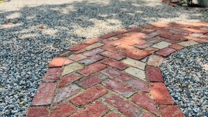 Un chemin de gravier, dont le revêtement est constitué de briques disposées en motifs, mène à un bâtiment à la façade en briques et doté d’une fenêtre.