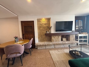 A modern living room with a stone fireplace, a flat-screen TV mounted above it, a round dining table with chairs, and a glass coffee table. - Nogalera- Apartamento reformado (Torremolinos)