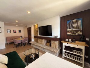 A living area with a sofa, dining table, chairs, a TV mounted on a stone wall, a coffee machine, and a bookshelf. - Nogalera- Apartamento reformado (Torremolinos)