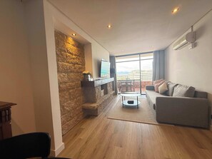 A modern living room with a stone accent wall, a flat-screen TV, a grey sofa, and a view of the cityscape through large windows.