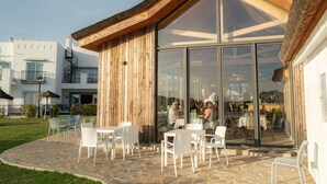 An outdoor dining area with stone flooring and white chairs, a wooden wall, and large glass windows.