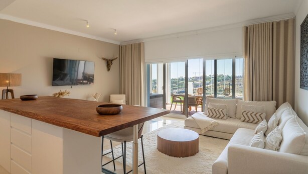 A modern living room with a wooden dining table, a sofa, and a large window offering a city view.