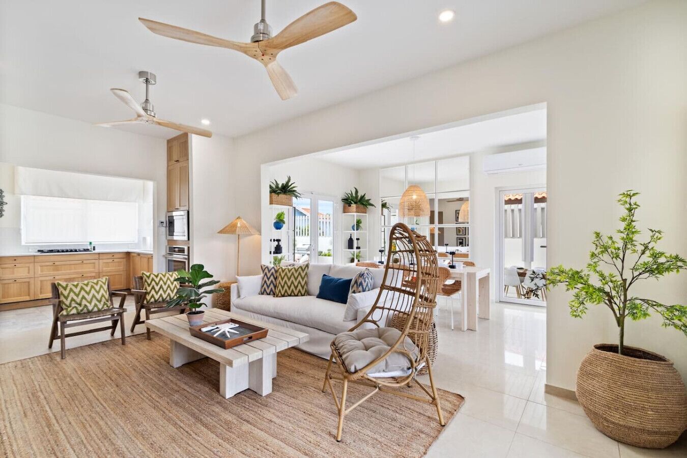 A spacious living room with a sofa, coffee table, and a wicker chair. There is a ceiling fan, a large window, and a kitchen area in the background.