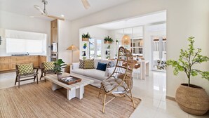 A spacious living room with a sofa, coffee table, and a wicker chair. There is a ceiling fan, a large window, and a kitchen area in the background. - 3BR Luxe Retreat with Private Pool by Bocobay (Noord)