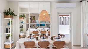 A dining area with a white table set for four, surrounded by wicker chairs, a hanging rattan light fixture, and shelves with decorative items and plants. - 3BR Luxe Retreat with Private Pool by Bocobay (Noord)