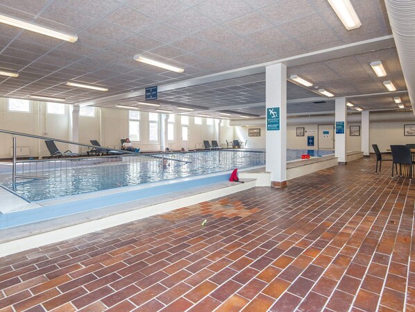 En inomhuspool med ett kaklat golv, ett genomskinligt tak med belysning och några personer som simmar.