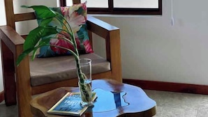 Una silla de madera con cojín y almohada estampada, una mesita de madera con un libro y un jarrón con una planta, y una ventana con vistas al océano y edificios.