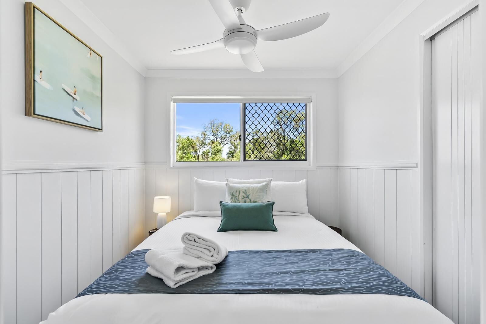 Un dormitorio con cama, almohadas, una toalla doblada, una mesita de noche, una lámpara, un ventilador de techo y una ventana con vista a árboles.
