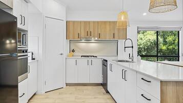 Eine moderne Küche mit weißen Schränken, einem Edelstahl-Kühlschrank, einem Einbauofen, einem Gasherd und einem großen Fenster mit Blick auf Grünflächen.