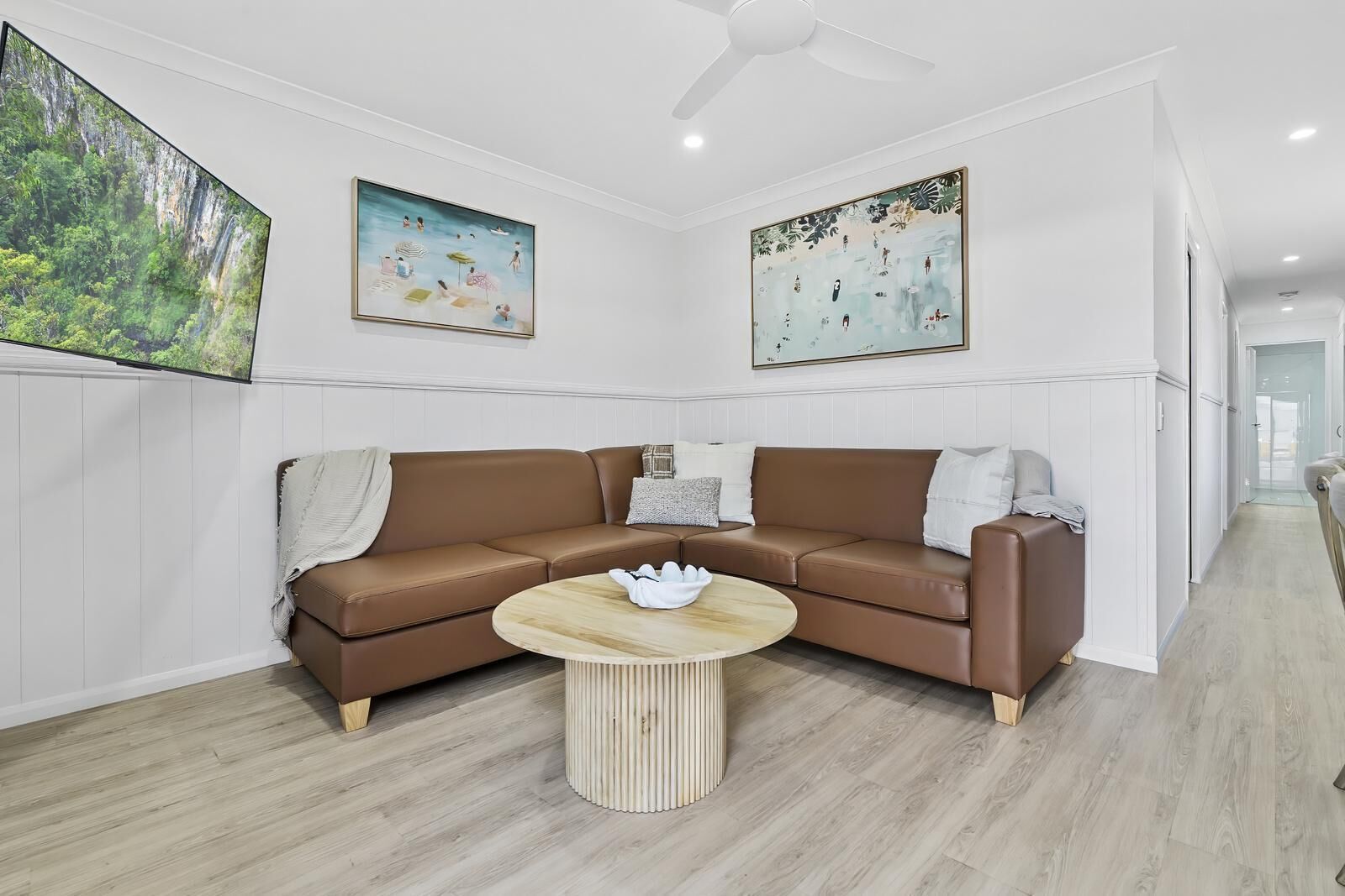 Uma sala de estar moderna com um sofá de couro marrom em formato de L, uma mesa de centro de madeira redonda e duas pinturas penduradas na parede.