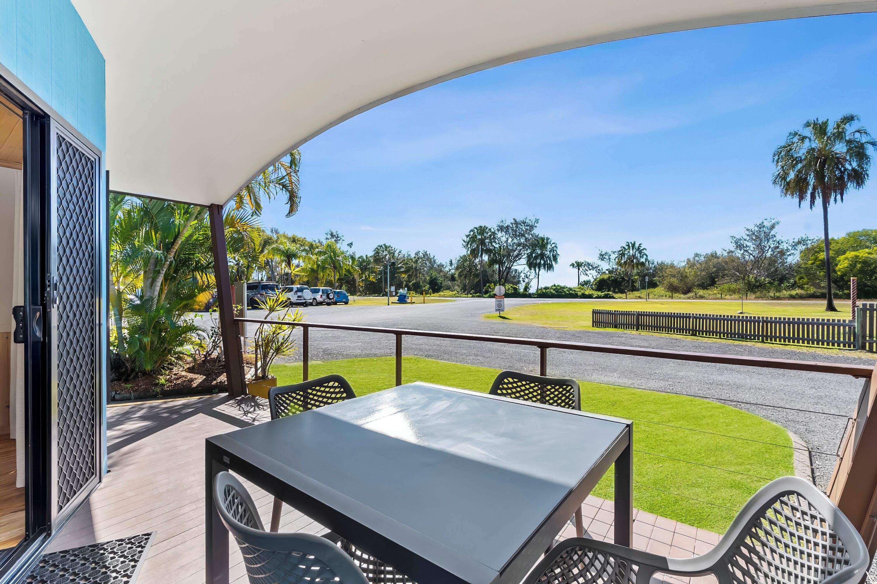 Un patio techado al aire libre con una mesa y sillas, vista a una carretera con vehículos y palmeras al fondo.