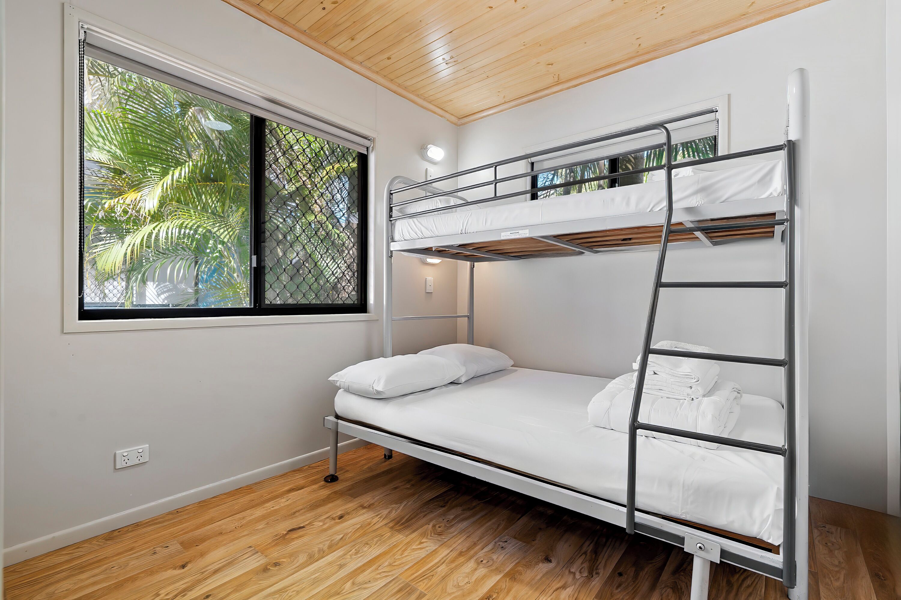 Een stapelbed met witte beddengoed, een raam met uitzicht op palmbomen en een houten plafond.