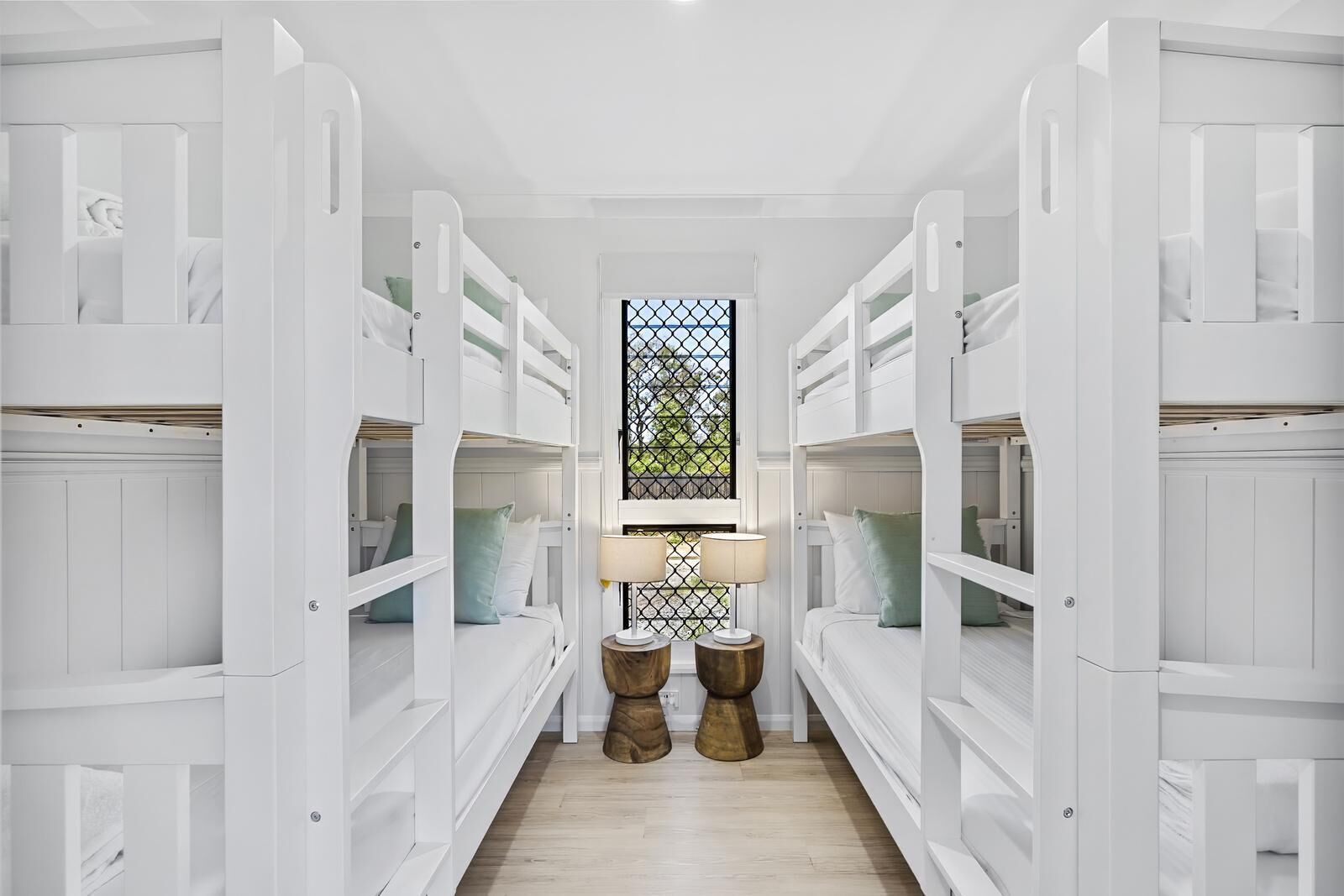 Un pasillo con literas, una ventana con celosía metálica y dos taburetes de madera.
