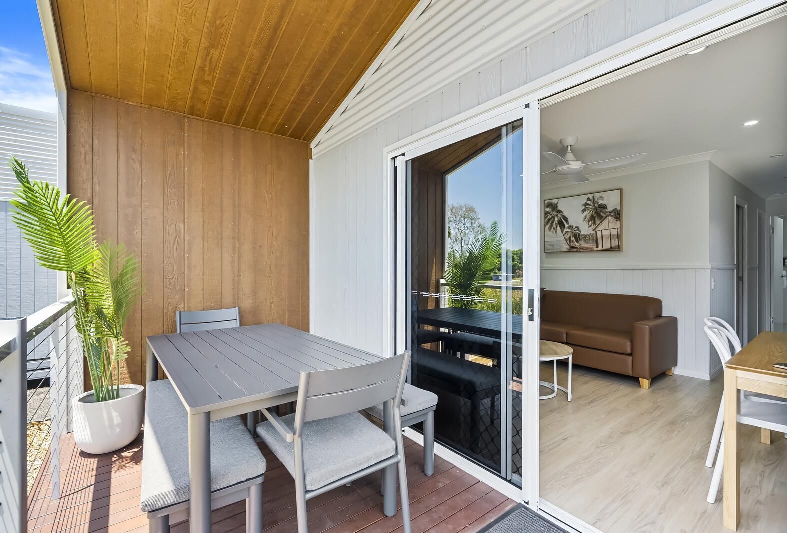 Un patio moderno al aire libre con techo de madera, una mesa de comedor con sillas, una planta en maceta y un ventanal corredero que da a una sala interior con sofá y un cuadro.