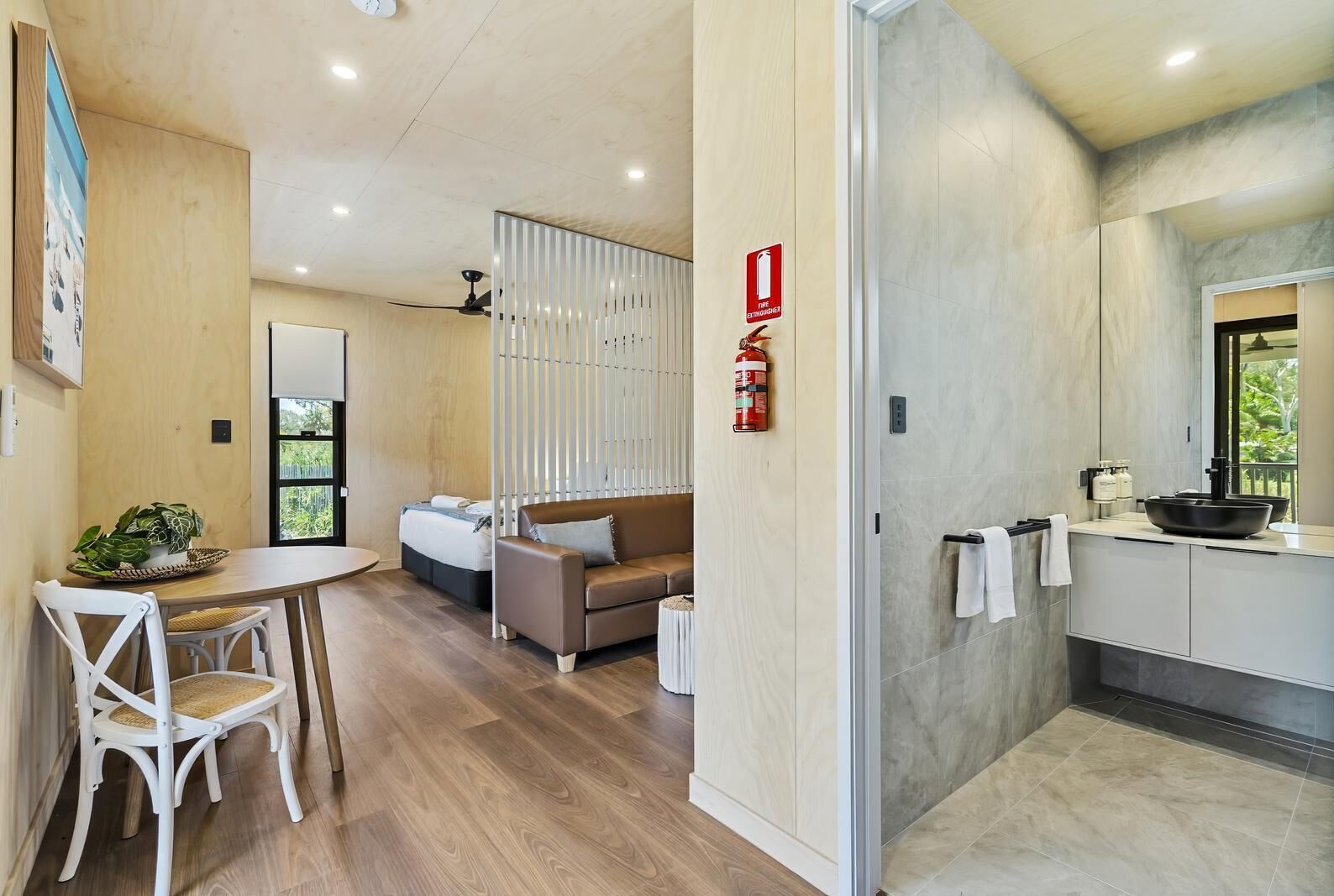 A modern hotel room with a wooden dining table, a sofa, a small kitchen area with a sink and a large window.