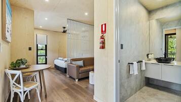 A modern hotel room with a wooden dining table, a sofa, a small kitchen area with a sink and a large window.