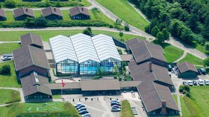 An aerial view of a building complex with a central swimming pool, surrounded by multiple smaller buildings, a parking area, and green lawns.