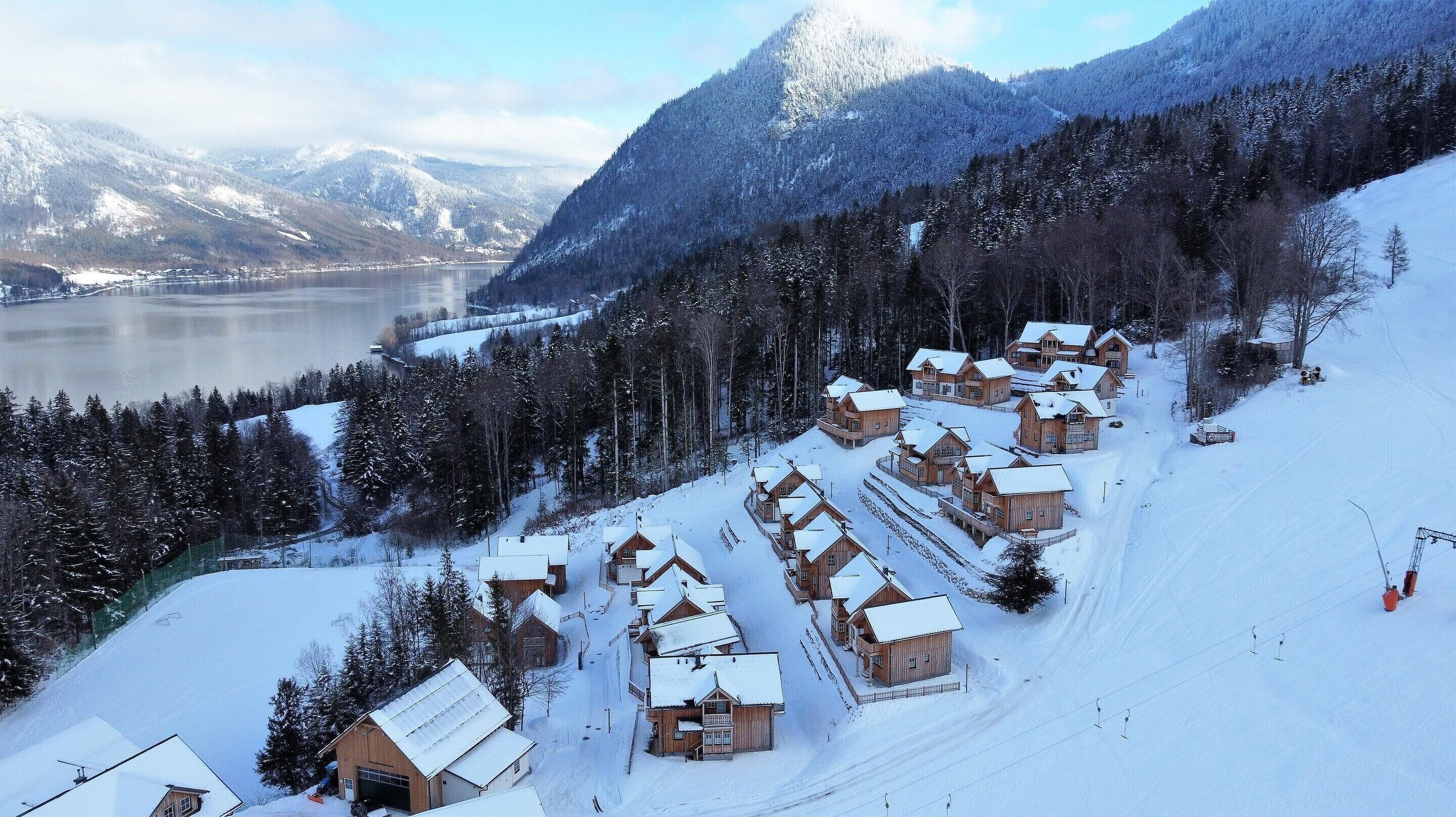A snow-covered mountain resort with wooden cabins, a ski slope, and a lake in the background.