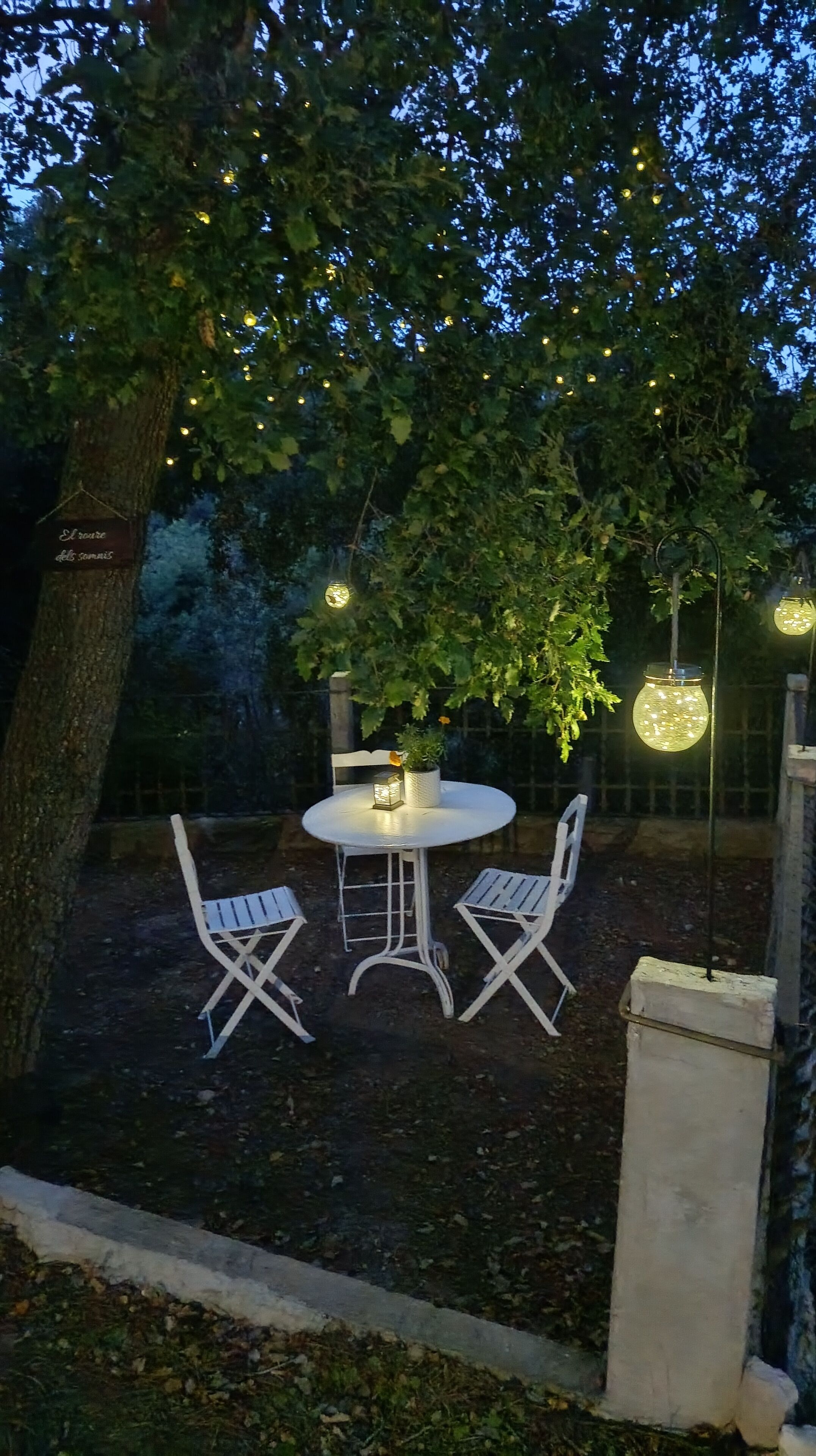 An outdoor seating area with a round table and two chairs, surrounded by trees and string lights.